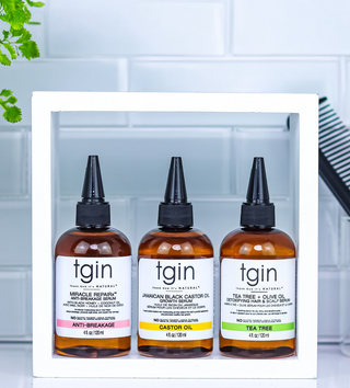 Three amber bottles of tgin haircare products displayed inside a white rectangular frame against a light tiled background. The bottles are labeled Miracle Reparative, Jamaican Black Castor Oil Conditioner, and Tea Tree & Olive Oil Conditioning Hair & Scalp Serum.